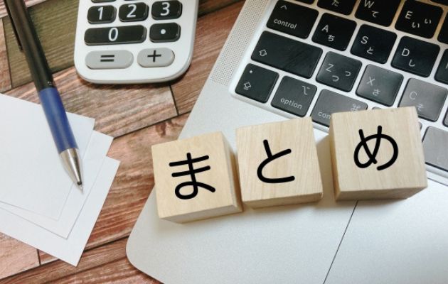 パソコンのキーボードの上に木のブロックで「まとめ」と書かれており、メモ紙や電卓、ボールペンが置いてある写真