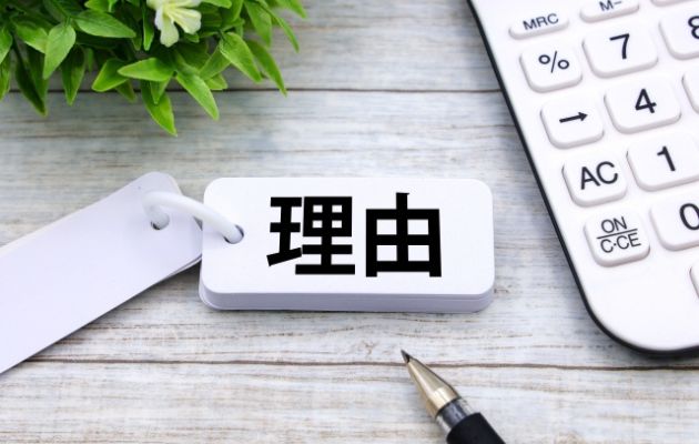 暗記カードに「理由」と書いてあり、周りに電卓とボールペン、緑の植物が置いてある写真